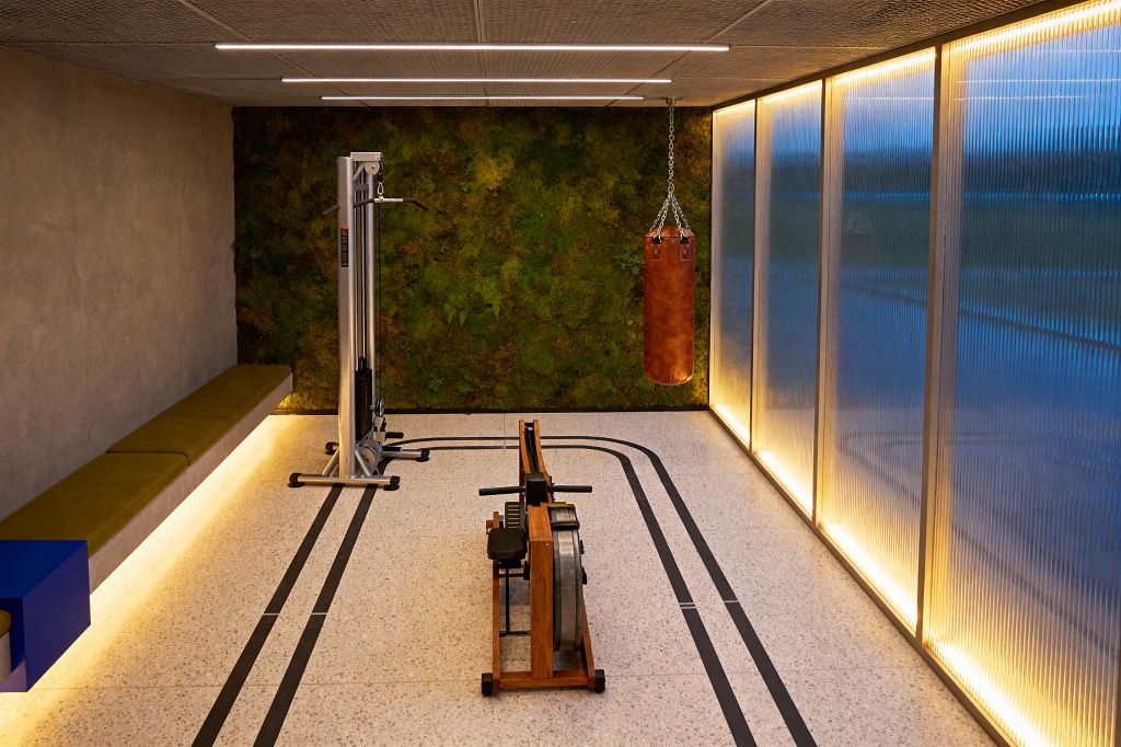 A long, minimalist home gym features a vibrant green moss wall at the far end, a wooden water rower centered on a speckled off-white terrazzo floor with black track lines, and a suspended cognac leather punching bag. The left wall is warm gray plaster with a floating olive bench lit from below, while the right side has full-height translucent panels washed in amber LEDs. Slim linear lights punctuate a textured acoustic ceiling.