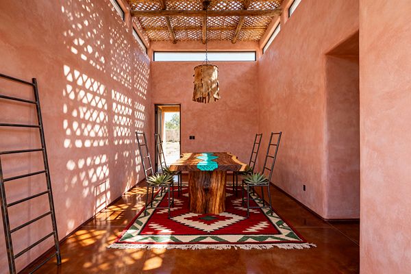 An elongated Southwestern dining room with adobe-pink plaster walls and a lattice wood ceiling casts patterned light across polished brown concrete floors, where a live-edge mesquite table with a turquoise inlay sits on a red and ivory kilim rug, surrounded by black iron ladder-back chairs. A rawhide fringe pendant hangs centered, clerestory windows line the upper walls, and an open door frames a desert landscape.