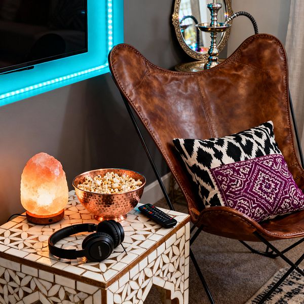 A cozy media room corner shows a camel-brown leather butterfly chair with black-and-white ikat and plum patterned cushions beside a white bone-inlay side table. On the table sit a glowing Himalayan salt lamp, a hammered copper bowl filled with popcorn, a TV remote, and black over-ear headphones, while teal LED light frames a wall-mounted screen; an ornate mirror and a metal hookah glint in the background against gray walls and a neutral rug.