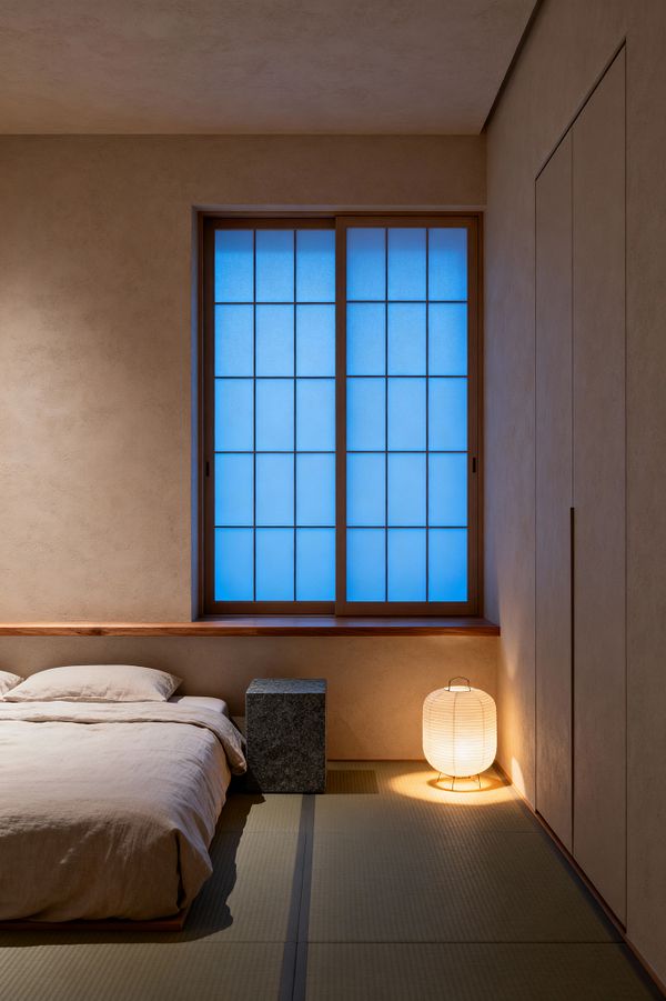 A minimalist bedroom with Japanese influence features a low bed dressed in beige linen beside a dark stone block nightstand. A large shoji window glows blue, framed in natural wood above a slim ledge. Warm plaster walls and built-in flush cabinets keep lines clean. Tatami mats span the floor, and a small paper lantern on metal legs casts a soft amber pool of light, creating a calm, uncluttered atmosphere.