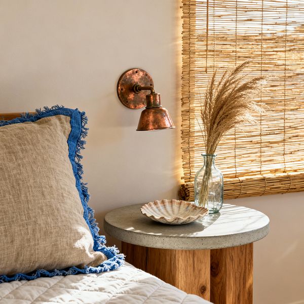 Close-up of a coastal-style bedside scene with a beige linen pillow edged in bright blue fringe on a white quilted bed. A round nightstand with a concrete top and wood base holds a clear glass bottle of dried grasses and a shell-shaped dish. An aged copper wall sconce is mounted on a warm white wall, while a rolled bamboo shade filters sunlight across the textures, creating a natural, relaxed look.