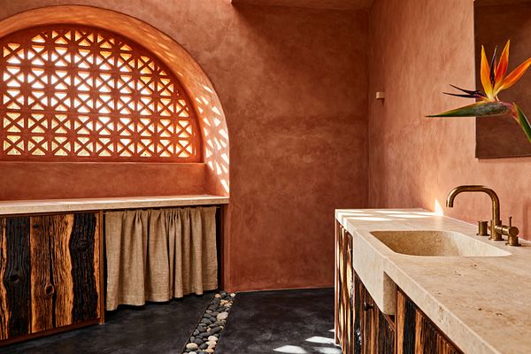 A biophilic laundry room features terracotta plaster walls and a large arched lattice window that casts patterned sunlight across the space. A pale stone countertop with an integrated sink and brass faucet pairs with rustic cabinetry in charred wood and a natural linen skirt; dark concrete floors include a pebble-lined channel, while a bird-of-paradise stem brings a vivid botanical note.