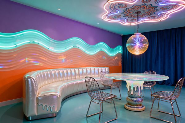 A colorful dining room features a curved pearlescent banquette against purple and orange walls, with backlit aqua acrylic waves casting a shifting glow, and a round iridescent resin table on a dripping pedestal surrounded by four chrome wire chairs. Overhead, a neon‑trimmed mandala ceiling frames a spherical plasma‑light pendant, while a teal floor and deep blue velvet curtain complete the retro‑futurist, psychedelic atmosphere.