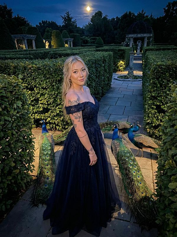 A woman in a dark gown poses in a beautifully landscaped garden with peacocks and a full moon in the background.