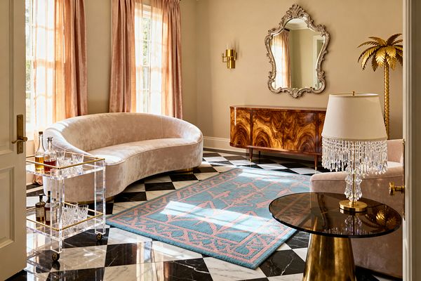 A vintage glam living room features a curved blush velvet sofa on a glossy black-and-white checkerboard marble floor, softened by a sky-blue rug with pink pattern. Champagne walls frame tall windows with pink velvet curtains. An ornate silver mirror hangs above a bookmatched walnut burl credenza beside a brass sconce. A gold palm floor light, acrylic bar cart with glassware, and a crystal-fringe table lamp add sparkle in warm natural light.
