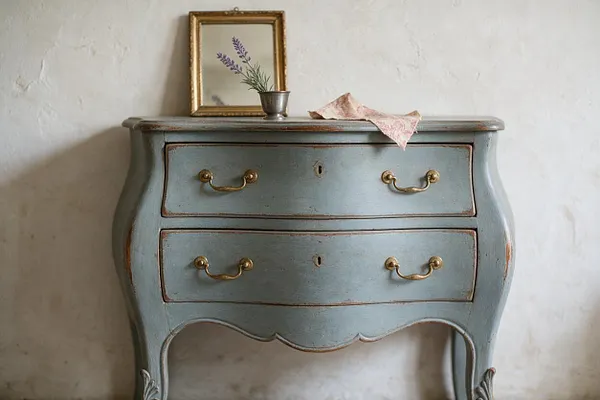 A distressed blue-gray bombe chest with curved sides and two drawers sits against a white plaster wall. Brass drawer pulls, worn edges, a small gilded frame, and a lavender sprig add to the soft French Country styling.