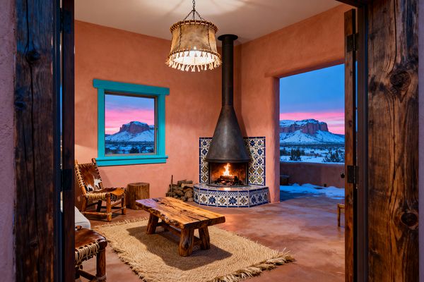 A Southwestern living room with terracotta adobe walls and turquoise window trim centers on a corner fireplace with a blackened steel hood and a blue-and-white Talavera tile surround. A live-edge wood coffee table sits on a braided jute rug between rustic cowhide chairs. A rawhide drum pendant hangs above, and stained concrete floors and stacked firewood add to the rustic feel. Large openings frame snowy desert mesas at sunset, mixing warm firelight with cool twilight colors.