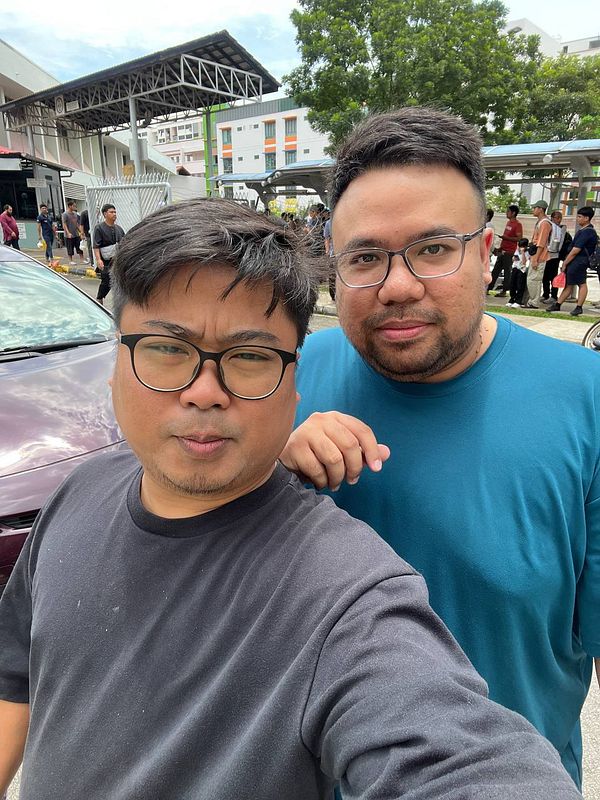 Two friends take a selfie outdoors with a lively background.