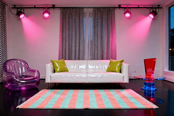 A Pop Art living room centers on a glossy white sofa with two lime pillows, set before windows draped in black-and-white polka-dot curtains. Magenta spotlights on ceiling tracks cast a neon wash on white walls. A translucent purple inflatable armchair sits on the left, and a clear acrylic side table with red and blue elements stands on the right. A mint and coral striped shag rug anchors a high-gloss black floor, creating a playful, retro-lounge feel.