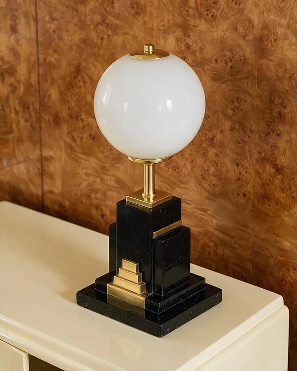A decorative table lamp with a frosted white globe shade, polished brass stem, and stepped black marble base sits on a cream-colored cabinet. The background features a rich burl wood wall panel, reinforcing the lamp’s glamorous Art Deco style.