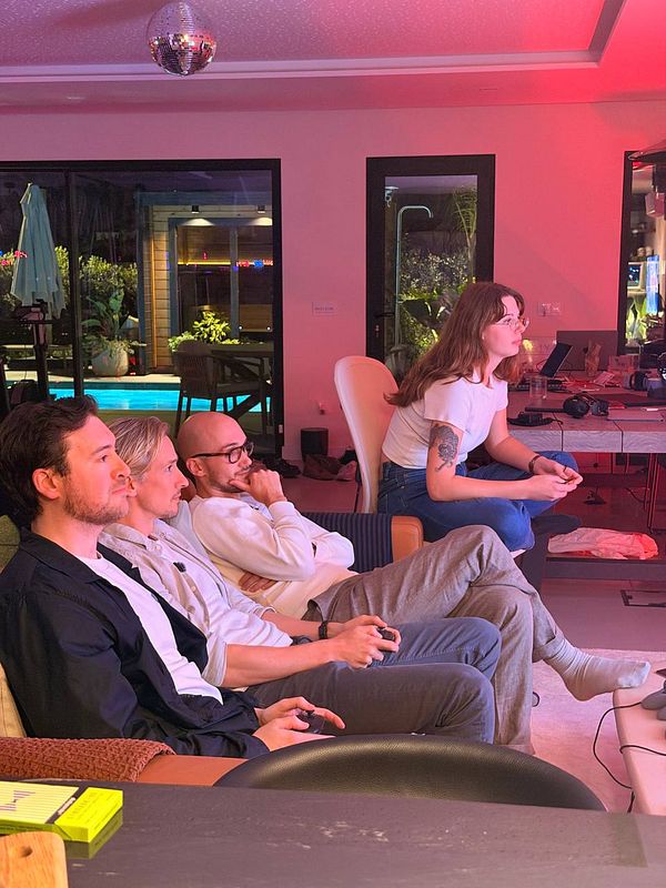 Four people engaged in an indoor gaming or coworking session in a room lit with pink and warm lighting near a poolside view.