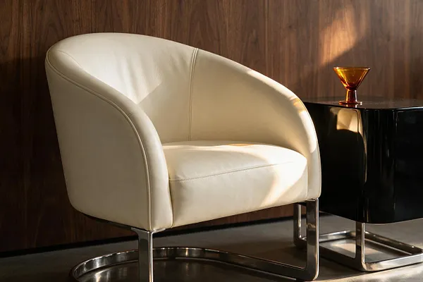 A cream upholstered tub chair with curved arms and a low, rounded back sits on a polished chrome sled base. Beside it is a glossy black side table with a small amber glass accent, set against a warm wood-paneled wall and lit by soft natural light.