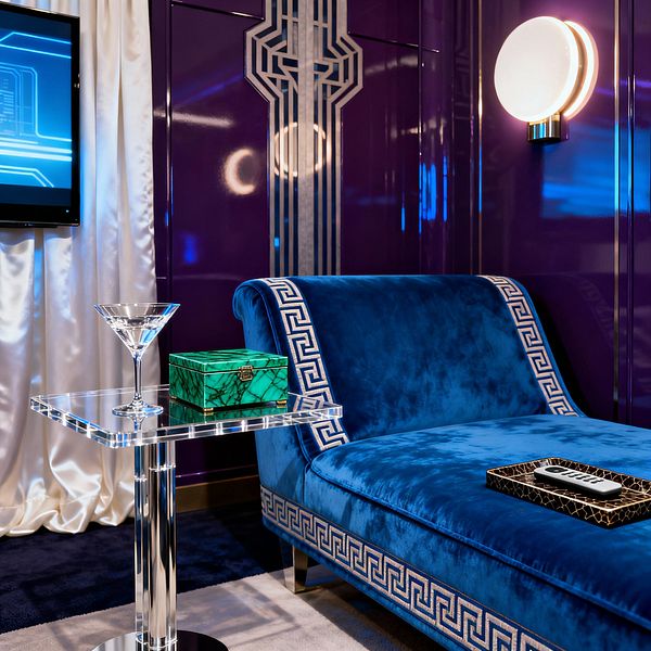 A jewel-toned media room showcases a sapphire blue velvet chaise with Greek key trim in front of glossy aubergine wall panels featuring Art Deco metallic inlay. A clear acrylic side table holds a martini glass and a small malachite box, while an ivory satin curtain frames a wall-mounted screen. A circular brass-and-opal sconce casts warm light over a deep navy rug, creating a glamorous Hollywood Regency setting for lounging and viewing.