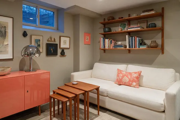 A cozy eclectic basement seating area with a white sofa, coral accent pillow, stacked wooden nesting tables, and a glossy coral cabinet topped by a chrome lamp. Floating wood shelves hold books and pottery above the sofa, while a small basement window and a gallery of framed art add warmth and personality.