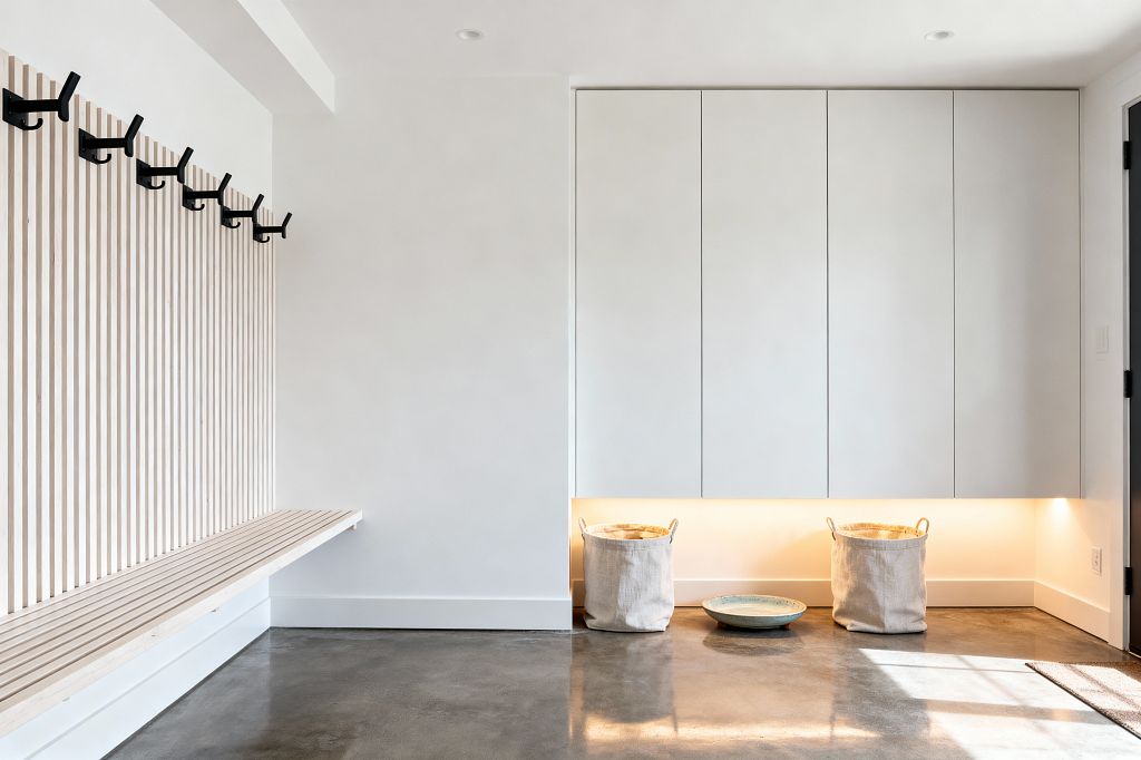 A minimalist mudroom with a pale wood slat wall and a floating slatted bench runs along the left, lined with matte black hooks. Opposite, flat-front light gray cabinets span the wall with a recessed toe-kick lit by warm LED, illuminating two off-white canvas baskets and a shallow ceramic tray on a polished gray concrete floor. Walls and ceiling are white with subtle recessed lights.