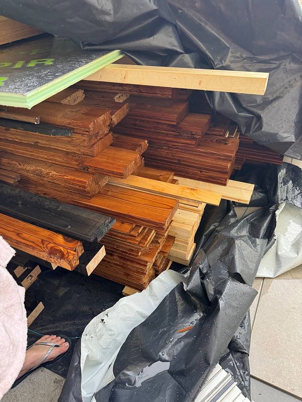 A stack of sauna wood is seen in a garden, partially covered by a black tarp, with some pieces exposed to rain.