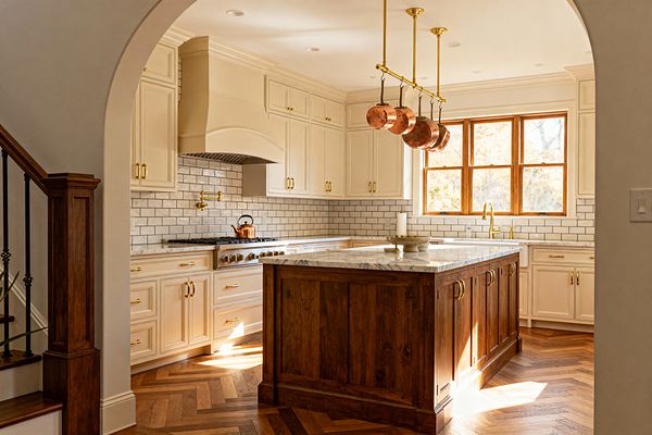 A traditional kitchen features cream inset cabinets with crown molding, brass hardware, and a marble-topped walnut island beneath a brass ceiling-mounted pot rack holding copper pans. A glossy white subway tile backsplash with dark grout runs around a stainless gas range with a brass pot filler, while herringbone wood floors, a wood-framed window, recessed lighting, and a brass bridge faucet complete the warm, classic look.