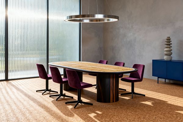 A contemporary dining room features an oval table with a light wood top, rounded metal rim, and two dark wood cylindrical bases, surrounded by six plum upholstered chairs on black four-star bases. Sunlight filters through floor-to-ceiling ribbed glass onto a warm cork floor, while a brushed steel ring pendant hangs above and a cobalt blue credenza with a stacked stone sculpture sits against soft gray plaster walls.