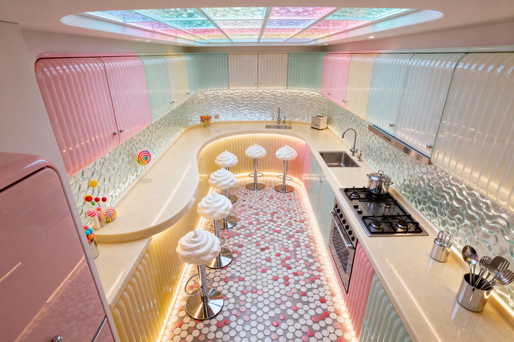A narrow U-shaped kitchen styled like Candy Land features glossy ribbed cabinets in pastel pink, mint, and butter yellow, cream solid-surface countertops, and a wavy textured glass backsplash. A curved breakfast bar seats five on white swirl-topped stools with chrome bases. The ceiling glows through multicolor backlit panels, while warm LED strips outline counters and toe-kicks. The floor is a penny tile mosaic in white, gray, and pink with lollipop decor on the counter.