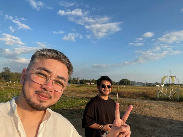 Two friends pose for a selfie in a scenic farmland setting under a clear blue sky.