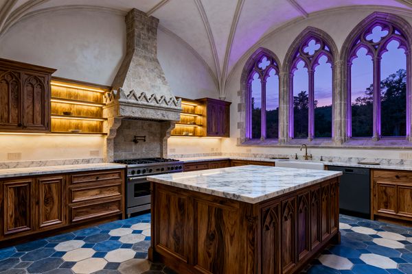 A Gothic-style kitchen features a ribbed vaulted ceiling, a carved stone range hood over a stainless professional range, and warm backlit open wood shelves flanked by walnut cabinets with pointed-arch detailing. A thick marble-topped island matches the perimeter counters above a navy, slate, and cream hexagon tile floor, while tracery arched windows glow violet at dusk beside a white apron-front sink with a brass faucet.