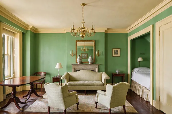 A traditional studio apartment with green walls, cream trim, and a polished wood floor. The room features a brass chandelier, a central fireplace with a large mirror, a cream settee, two upholstered armchairs, a patterned area rug, a wooden dining table by the window, and a bed tucked into an alcove on the right.