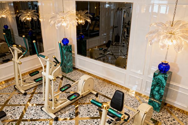 A glamorous home gym features cream cardio machines with gold hardware, teal grips, and peacock-print footplates on a terrazzo floor inlaid with brass strips. Glossy white paneled walls hold large gold-trimmed mirrors, while malachite-pattern pedestals with cobalt-blue glass bases support palm-frond crystal pendant lights that cast warm illumination. The symmetrical, reflective setting shows additional exercise equipment in the mirror, emphasizing jewel tones and metallic finishes.
