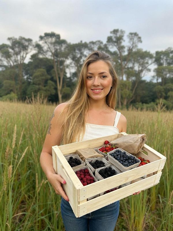 A woman holds a wooden crate filled with various berries in a field.