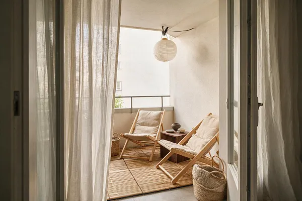 A small Japandi-style patio with two light wood lounge chairs and cream cushions arranged around a low wooden table. Sheer linen curtains frame the view, while a paper lantern, woven baskets, and a bamboo mat add soft natural textures in a warm neutral palette.
