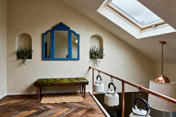A compact, loft-style home gym with limewashed plaster walls and a sloped ceiling lit by a skylight. A cobalt-blue trifold mirror hangs above an olive green tufted leather bench on a jute rug over herringbone oak flooring. Arched wall niches hold greenery, and a copper pipe balustrade displays vintage white enamel kettles used as weights. A copper pendant with a linen shade adds warm light to the rustic, elegant space.