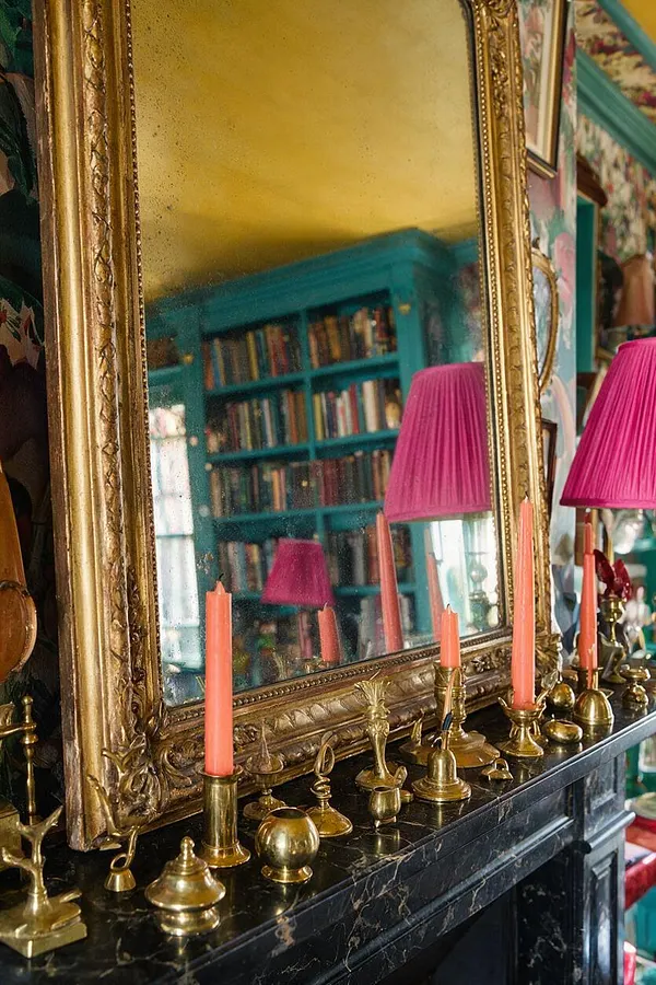 A large ornate gold-framed mirror rests above a black marble mantel covered with assorted brass decorative objects and coral candles. In the reflection, a turquoise bookcase, a yellow ceiling, and a bright pink lampshade reveal a richly layered maximalist room.