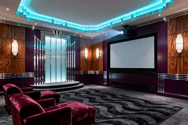 A luxurious Art Deco home theater with deep plum walls, walnut veneer panels, and polished chrome trim. A stepped ceiling with turquoise cove lighting surrounds the room. At the front, a backlit portal of frosted glass panels framed by mirror-like fins sits on curved steps. White geometric sconces flank a wide projection screen with black speakers. Plush burgundy velvet lounge chairs face a gray carpet with black wave pattern.