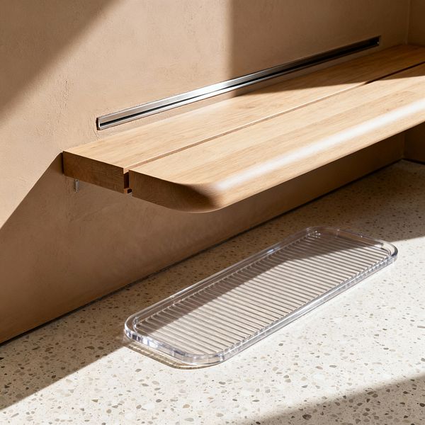 A minimalist mudroom corner featuring a floating light-oak bench with a rounded front edge, mounted to a smooth beige plaster wall with a slim recessed stainless-steel rail. Below, pale speckled terrazzo flooring is lit by sunlight, and a clear ribbed acrylic tray sits ready for wet shoes. Clean lines, warm neutrals, and hidden brackets create a calm, functional, clutter-free entry space.