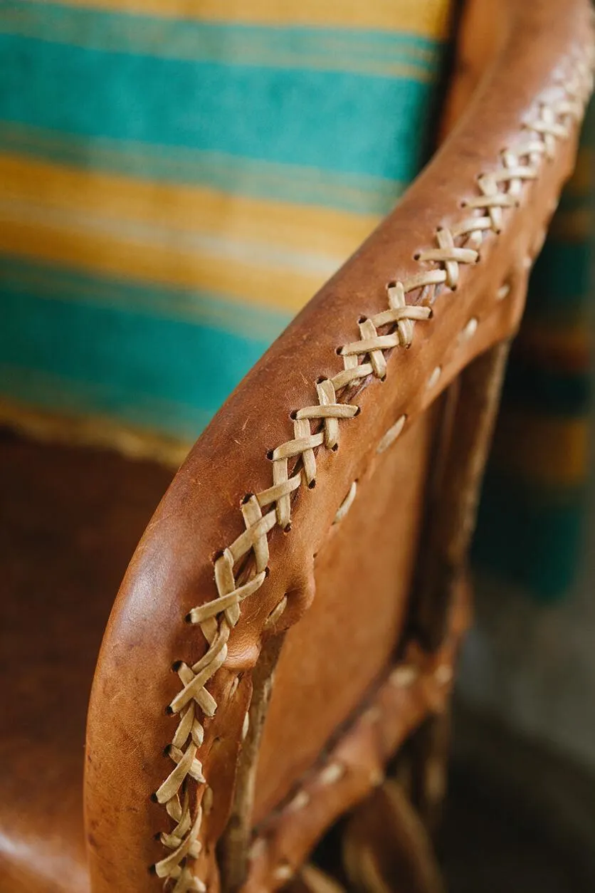Close-up of a Southwestern-style leather chair with a rounded armrest stitched together using visible rawhide lacing. A turquoise and gold striped textile appears softly blurred in the background, adding color and warmth.