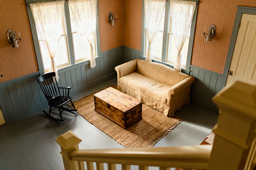 View from a staircase into a cozy farmhouse living room with blue-gray beadboard wainscoting and warm peach beadboard above. Two sets of windows with sheer lace curtains brighten a tan slipcovered loveseat and a rustic wood trunk used as a coffee table on a jute rug. A black rocking chair sits to the left over slate-gray painted plank floors. Metal candle sconces and a white paneled door complete the vintage look.