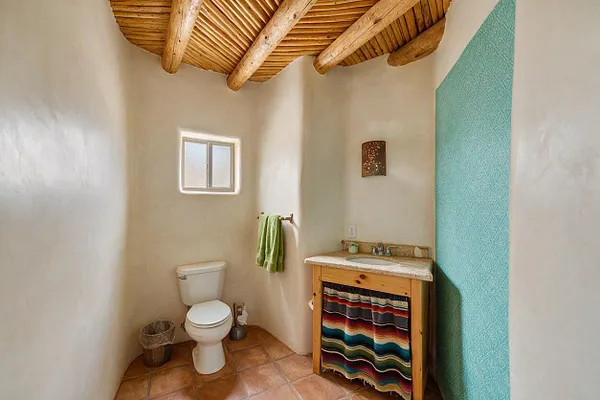 A compact Southwestern powder room with smooth adobe-style walls, a rustic wood-beam ceiling, terracotta tile flooring, and a small frosted window. The space includes a white toilet, a natural wood vanity with a stone countertop, a turquoise patterned accent wall, and a colorful woven fabric curtain below the sink.