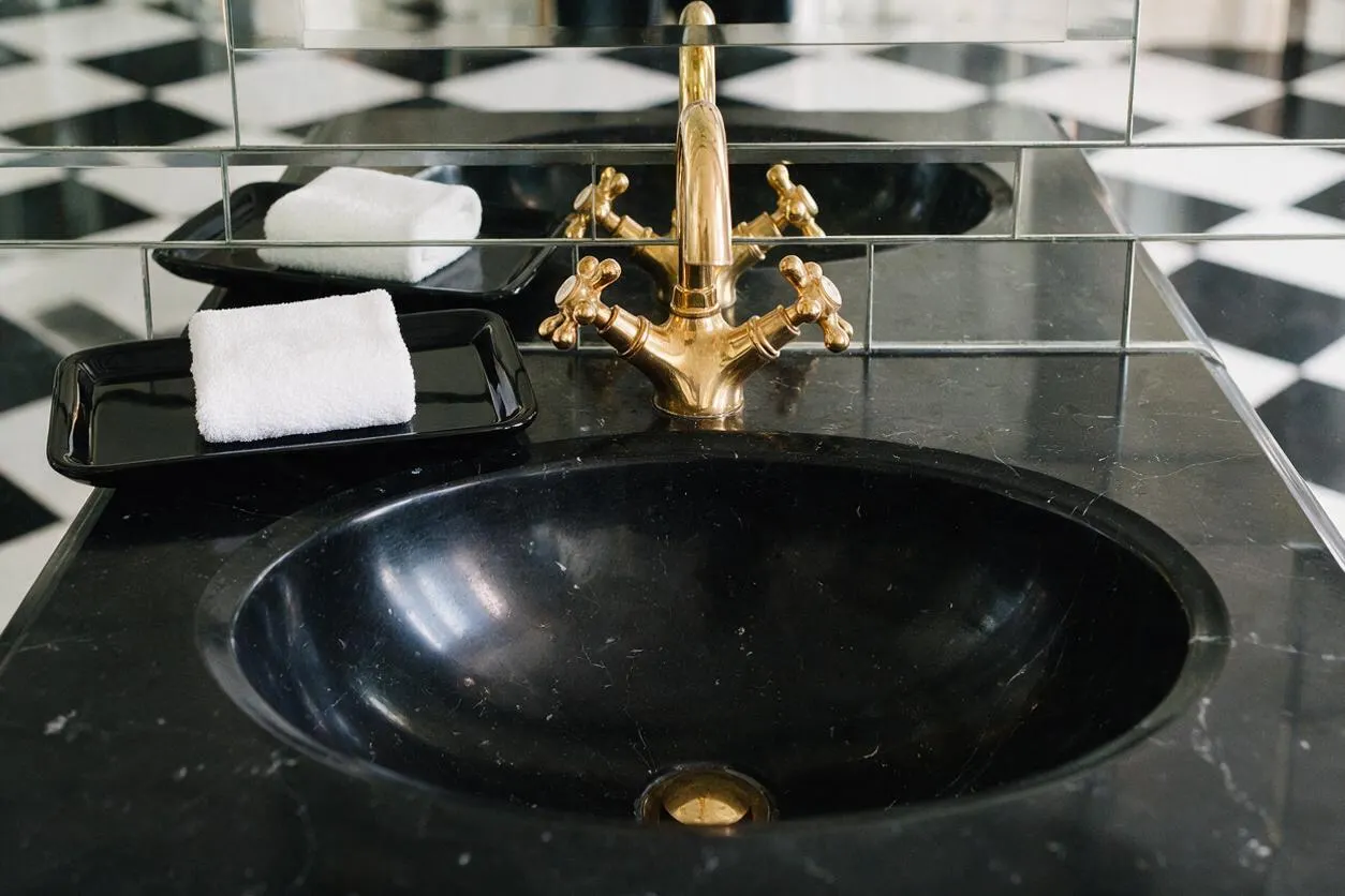 A bathroom vanity detail with a black marble countertop and integrated black sink, paired with a polished brass faucet. Behind it, mirrored wall tiles reflect the space, and a black-and-white checkerboard floor adds a bold graphic pattern. Two black trays with rolled white towels sit beside the sink.