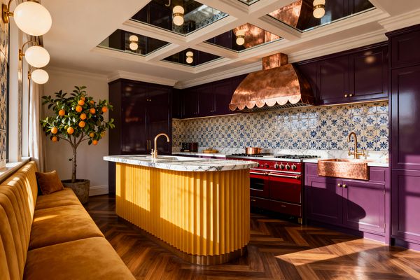 A vibrant kitchen features aubergine cabinets and a burgundy range beneath a scalloped copper hood, set against a blue-and-white patterned tile backsplash. A marigold fluted island with a veined marble top and brass faucet sits on a walnut herringbone floor, while a copper apron sink, ochre velvet banquette, globe sconces, mirrored coffered ceiling, and a potted orange tree complete the richly layered space.