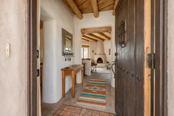 A Southwestern entryway with thick white adobe walls, exposed wooden ceiling beams, and a heavy dark wood front door. A carved wood console table, decorative mirror, and colorful woven runner add warmth, while a view into the living area reveals a kiva fireplace and rustic seating.