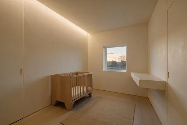 A sparse minimalist nursery with smooth beige walls, a natural oak crib with vertical slats, and a neutral rug on the floor. A small window brings in soft sunset light, and a floating wall-mounted shelf adds a simple functional detail.