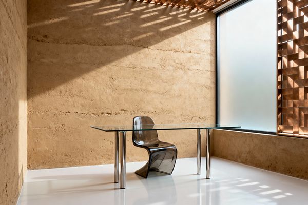 A minimalist home office features a rectangular glass desk with polished cylindrical steel legs and a smoked acrylic cantilever chair. Warm rammed-earth walls meet a large frosted window screened by a bronze perforated lattice that continues across the ceiling, casting geometric shadows on a glossy white floor; the uncluttered space emphasizes clean lines, natural texture, and filtered daylight.