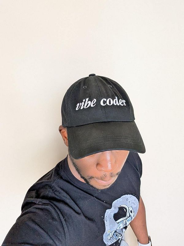 A person wearing a black cap with the text 'vibe coder' is shown against a plain background.
