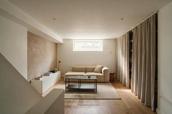 A modern basement lounge with a beige two-seat sofa centered against the back wall, a low glass-and-black metal coffee table, and a textured rug over light wood flooring. Soft floor-length curtains line the right side, while a small horizontal window brings in daylight above the sofa.