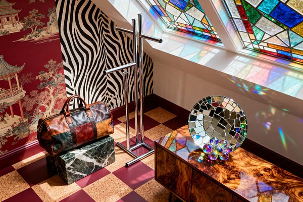 A maximalist home gym corner features black-and-white zebra wallpaper meeting deep burgundy chinoiserie, with a slanted skylight of multicolored stained glass casting jewel-toned reflections across the space. On a checkered floor of burgundy tiles and cork squares sit a chrome freestanding rack, a green marble plinth holding a patchwork leather duffel bag, and a glossy burl wood console topped with a round mosaic mirror and iridescent glass spheres.