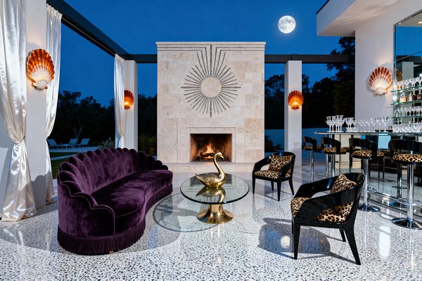 An open-air patio at night features a cream travertine fireplace with a sunburst motif, framed by white columns and satin drapes. A scalloped deep-purple velvet sofa with fringe faces a round glass table on a brass base topped by a gold swan. Two black chairs and bar stools upholstered in leopard print sit by a mirrored bar lined with crystal. Terrazzo flooring gleams under warm amber shell sconces, with the moon visible beyond.