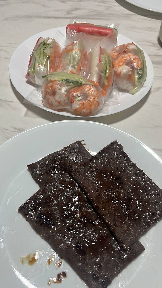 A close-up of a white plate containing a serving of Vietnamese spring rolls accompanied by a decorative sauce garnish and green leafy vegetables.