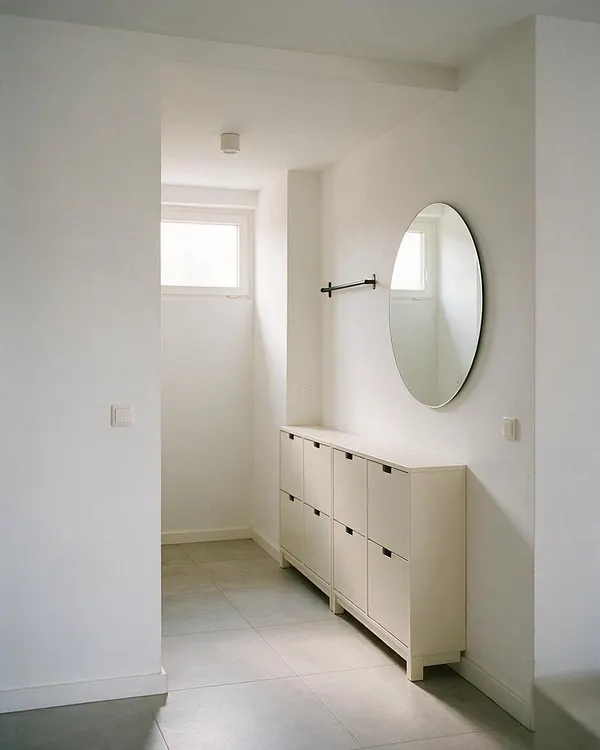 A bright minimalist entryway with white walls, a narrow cream-colored storage cabinet along the right wall, and a large oval wall mirror above it. The floor is light gray tile, and a small high window brings soft natural light into the simple, uncluttered space.