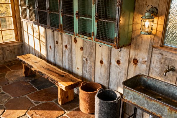 A rustic mudroom features weathered barn-wood walls, a live-edge wood bench, and vintage mint-green metal lockers with wire-mesh doors and hooks beneath. A galvanized trough sink with a wall-mounted faucet sits beside a caged lantern sconce, while felted wool baskets rest on a terracotta and charcoal flagstone floor; wood-framed windows and textured glass bring warm natural light across the space.