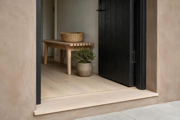 A minimalist Japandi-style entryway with beige plaster walls, a light wood threshold and floor, a black vertical-plank door, and a simple wooden bench holding a woven basket. A small potted plant in a muted ceramic pot adds softness to the calm, natural composition.