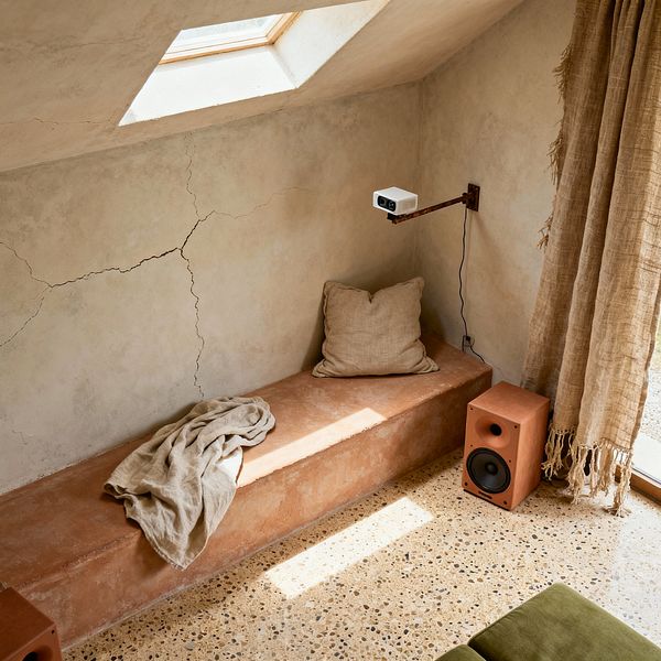 A small media room with limewash plaster walls showing natural cracks, a clay-rendered built-in bench topped with a neutral linen pillow and a casually draped throw, and speckled terrazzo flooring. A compact white projector is mounted on a rusted steel swing arm above a terracotta-colored speaker, while loosely woven, fringed natural drapes hang near a glass door; an olive cushion peeks in, all bathed in light from a roof skylight.